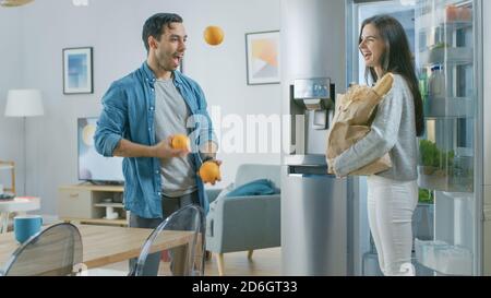 Schöne junge Paar haben Spaß in der Küche. Der Mensch jongelt mit Orangen. Mädchen klatscht, jubelt für ihn und sie lachen. Das Zimmer ist modern Stockfoto