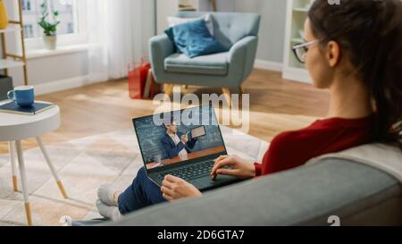 Junge Frau zu Hause verwendet Laptop für Video-Anruf mit seinem Lehrer. Bildschirm zeigt Online-Vortrag mit Lehrer erklären Thema. Er sitzt auf einer Couch Stockfoto