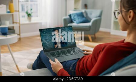 Junge Frau zu Hause verwendet Laptop für Video-Anruf mit seinem Lehrer. Bildschirm zeigt Online-Vortrag mit Lehrer erklären Thema. Er sitzt auf einer Couch Stockfoto