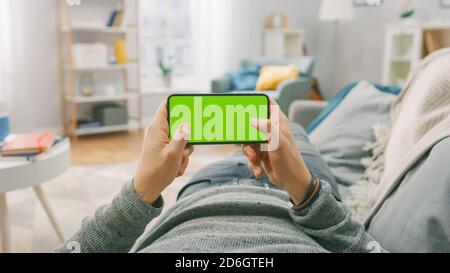 Der Mann zu Hause, der mit dem Smartphone auf einer Couch liegt, hält ihn horizontal im Landschafts-Modus. Videospiele Spielen, Videos Ansehen. Point-of-View-Aufnahme. Stockfoto