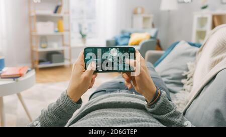 Der Mann zu Hause, der mit dem Smartphone auf einer Couch liegt, hält ihn horizontal im Landschafts-Modus. Er spielt First Person Shooter Video Game. Standpunkt Stockfoto