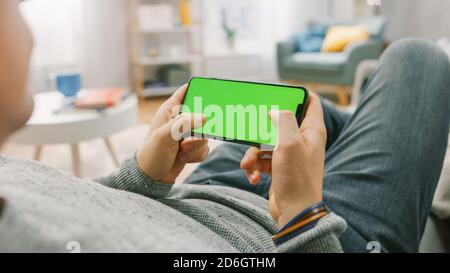 Der Mann zu Hause, der mit dem Smartphone auf einer Couch liegt, hält ihn horizontal im Landschafts-Modus. Tasten drücken, um Videospiel zu spielen. Kameraaufnahme vom Standpunkt aus. Stockfoto