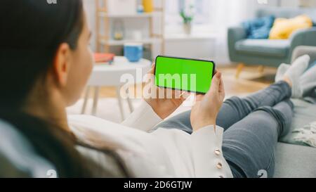 Frau zu Hause, die mit dem Smartphone auf einer Couch liegt, hält sie horizontal im Landschafts-Modus. Kameraaufnahme vom Standpunkt aus. Stockfoto