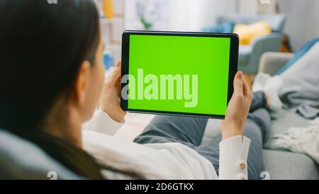 Junge Frau zu Hause ruht auf einer Couch mit Green Mock-up Screen Tablet-Computer im horizontalen Querformat. Frau mit Gesten mit Stockfoto