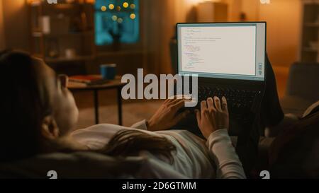 Abend zu Hause: Junge Programmiererin Frau auf einer Couch liegend mit Laptop-Computer zum Schreiben von Script Code. Smart Lady ist App Developer, die von zu Hause aus arbeitet Stockfoto