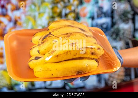 Neue Normalität nach der Covid-19 Pandemie: Eine Hand mit blauen Schutzhandschuhen hält eine Plastiktüte mit Bananen. Im Hintergrund Gemüse, Obst Stockfoto