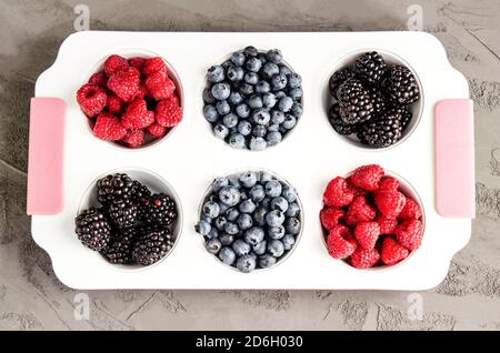 Frische Himbeeren, Blaubeeren und Brombeeren in einer weißen Eisenkalzform auf grauem Betongrund. Nahaufnahme, Saisonkonzept, Sommersaison. Er Stockfoto