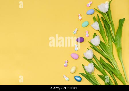 Ostereier farbige, dekorative Wäscheklammern für Dekor und ein Strauß weißer Tulpen auf gelbem Hintergrund, Kopierraum, flach liegend. Für Text platzieren. Der Stockfoto