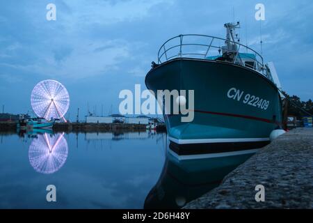 Das Boot schwimmt im Hafen in Honfleur während des bewölkten Abends. Das Riesenrad dreht sich hinter der Spiegelung im Wasser. Stockfoto