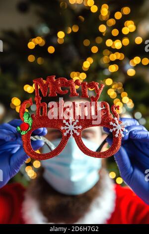Mann, der eine Brille mit Frohe Weihnachten hält und Schutzmaske und Handschuhe für Coronavirus, Covid-19 und Holiday Concept trägt Stockfoto