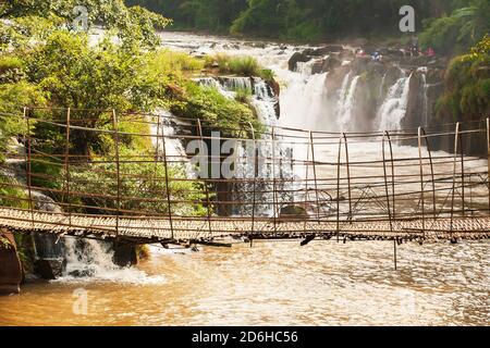 Eine ruhige alte Bambusfußbrücke über den TAD Pha Suam Wasserfall in der Dämmerung, laotische Familie, die sich im Hintergrund entspannt. Bolaven Plateau, Süd-Laos. Stockfoto