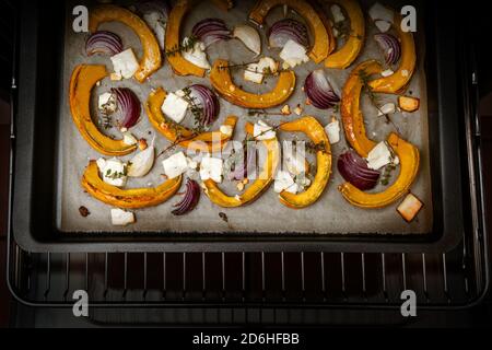 Kürbis- oder Kürbisscheiben mit roten Zwiebeln, Knoblauch, Feta-Käse und Thymian auf einem Backblech im Ofen, herbstliches Gemüsegericht für Thanksgiving und Hallo Stockfoto