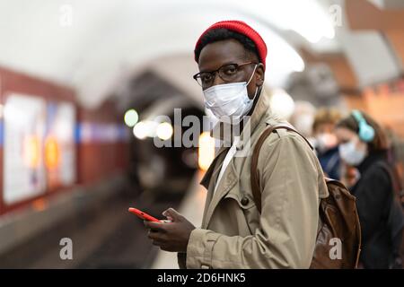 Afro-amerikanischer Millennial-Mann in Trenchcoat, roter Hut mit Gesichtsmaske als Schutz gegen covid-19, Grippe-Virus, warten auf den Zug in U-Bahn-stat Stockfoto