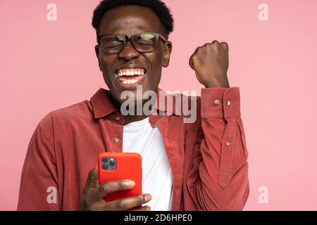 Aufgeregt afro-amerikanischen Millennial Mann in Brille halten Handy isoliert auf rosa Studio Hintergrund. Überglücklich schwarzer Mann Blick auf Smartphone, Lächeln fe Stockfoto