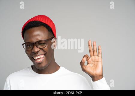 Fröhlich verspielt junge Afro-Amerikaner tragen roten Hut in guter Stimmung lächelnd breit, zwinkend, zeigt okay Geste über Studio grauen Hintergrund. Positiv Stockfoto