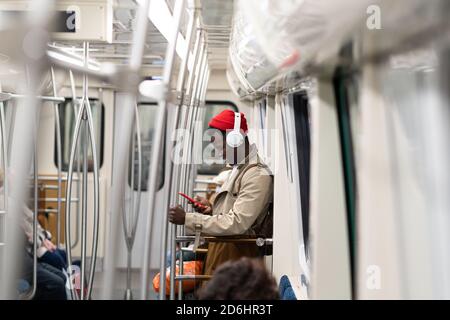 Afro-amerikanischer Passagiermann in rotem Hut, Trenchcoat Stand in U-Bahn-Zug, mit Handy, hört Musik mit drahtlosen Kopfhörern in öffentlichen tra Stockfoto