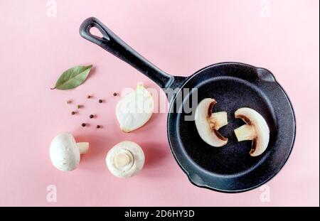 Kleine Pfanne aus schwarzem Gusseisen mit Champignon-Scheiben, Pilzen, Zwiebeln und Gewürzen auf rosa Oberfläche. Horizontale Ausrichtung, selektiver Fokus, Top Vie Stockfoto