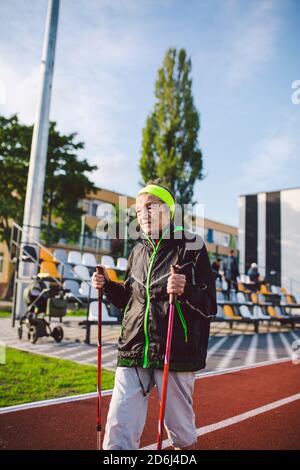 Aktive ältere kaukasische ältere Frauen von 90 Jahren üben Nordic Walking mit Skistöcken auf einer Strecke mit roter Gummibeschichtung. Aktivurlaub. Eine Passform Stockfoto