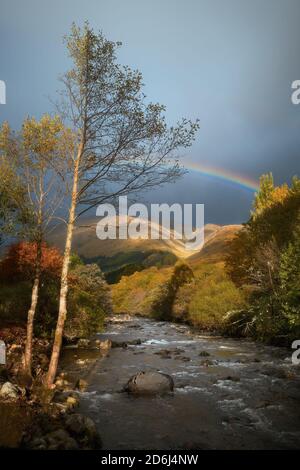 Bergfluss von Herbstbäumen nach Regen umgeben. Es gibt einen Regenbogen im wolkigen Himmel. Glenfinnan, Highlands, Schottland Stockfoto