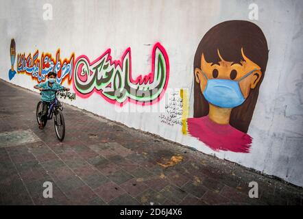 Ein palästinensischer Junge fährt mit dem Fahrrad an einem Wandgemälde vorbei, auf dem ein Kind mit einer Maske an einer Wand in Khan Yunis im Süden des Gazastreifens abgebildet ist. Stockfoto