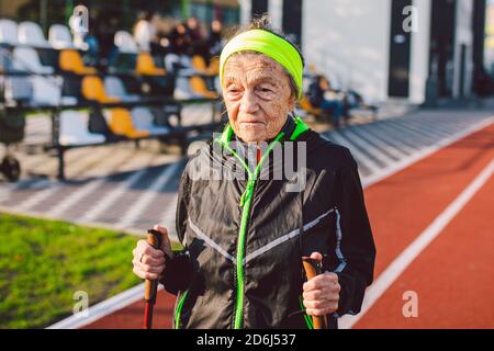Aktive ältere kaukasische ältere Frauen von 90 Jahren üben Nordic Walking mit Skistöcken auf einer Strecke mit roter Gummibeschichtung. Aktivurlaub. Eine Passform Stockfoto