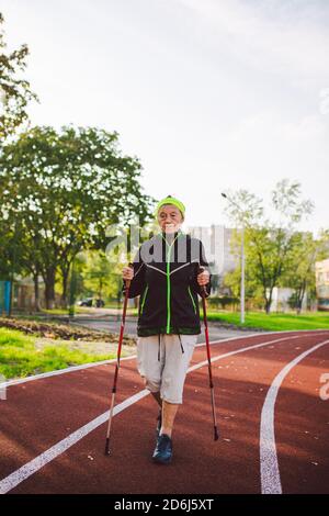 Ältere Frau, die mit Wanderstöcken im Stadion auf einer roten Gummihülle läuft. Ältere Frau 88 Jahre alt macht Nordic Walking Übungen in der Stadt Stockfoto