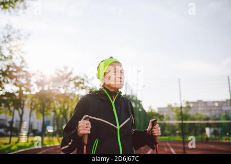 Ältere Frau, die mit Wanderstöcken im Stadion auf einer roten Gummihülle läuft. Ältere Frau 88 Jahre alt macht Nordic Walking Übungen in der Stadt Stockfoto