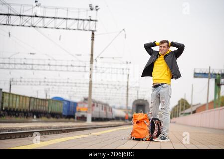 Der junge Mann steht auf dem Bahnsteig und wartet auf den Zug. Männlicher Passagier mit Rucksäcken auf Bahnsteig in Warten auf Zugfahrt. Konzept des Tourismus Stockfoto