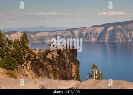 Am späten Nachmittag sitzt eine Frau am Rand einer Klippe, während eine Fotografin ihr Foto mit dem wunderschönen blauen Kratersee als Hintergrund macht. Stockfoto