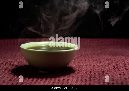 Chinesischer Tee dampfend in einer kleinen grünen Teetate isoliert mit Ein schwarzer Hintergrund und eine rote Oberfläche Stockfoto