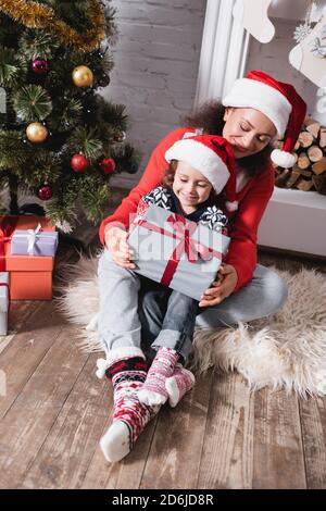 Mutter umarmt Tochter mit Geschenkbox neben festlichen Kiefer und Kamin Stockfoto