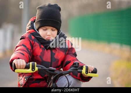 Trauriger, wütender Junge mit Hut und Schal, der auf dem Fahrrad im Park sitzt. Stockfoto