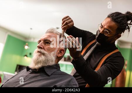 Der junge afroamerikanische Barbier und Dreadlocks mit einer Maske zur Prävention von Coronaviren kämmt mit einem Kamm und seinen Händen einen reifen weißen Kaukasier Stockfoto
