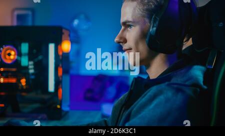 Glücklicher Gamer und Blogger in Headset mit einem Mikrofon spielen Shooter Online-Video-Spiel auf seinem PC. Raum und PC haben farbenfrohe Neon LED-Leuchten Stockfoto