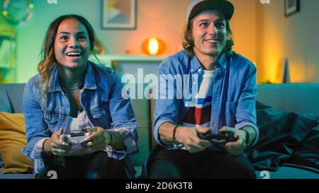 Aufgeregt Black Gamer Girl und Young man sitzen auf einer Couch und spielen Videospiele auf der Konsole. Sie spielen mit Wireless-Controllern. Gemütliches Zimmer ist beleuchtet Stockfoto