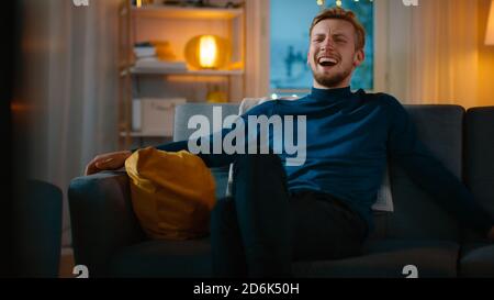 Am Abend Handsome Young man zu Hause sitzen auf einer Couch funktioniert auf einem Laptop-Computer, und Uhren TV, lacht bei Comedy. Stockfoto