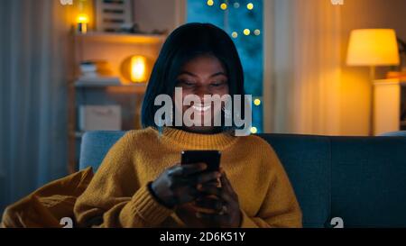 Portrait of Beautiful Black Girl nutzt Smartphone während Sie auf einer Couch zu Hause sitzen, ist ihr Gesicht mit einem Bildschirmlicht beleuchtet. In der niedlichen Frau Stockfoto