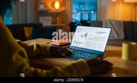 Laptop-Computer mit statistischen Infografiken steht auf einem Schreibtisch im Wohnzimmer. Im Hintergrund gemütliches Wohnzimmer mit warmen Licht an. Stockfoto