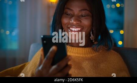 Portrait of Beautiful Black Girl nutzt Smartphone während Sie auf einer Couch zu Hause sitzen, ist ihr Gesicht mit einem Bildschirmlicht beleuchtet. In der niedlichen Frau Stockfoto