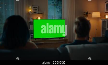Couple Watches Green Mock-up Screen TV, während Sie auf einer Couch im Wohnzimmer sitzen. Romantischer Abend für Freund und Freundin. Stockfoto