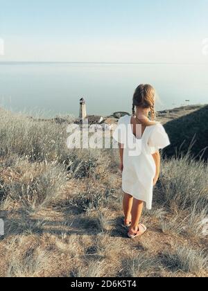 Ganzkörper-Rückansicht von einsamen unkenntlich Mädchen in weiß Sommerkleid steht auf grasbewachsenen Hügel an der Küste mit Leuchtturm Turm Stockfoto