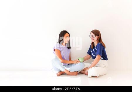 Volle Länge der glücklichen multirassischen weiblichen Jugendlichen in lässiger Kleidung Mit einer Tasse Getränk sitzend mit Beinen gegen Weiß gekreuzt Hintergrund und Chat Stockfoto