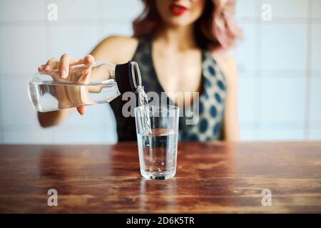 Crop schlank weiblich in stilvollem Outfit gießen frisches Wasser aus Transparente Flasche in Glas auf Holztisch platziert Stockfoto