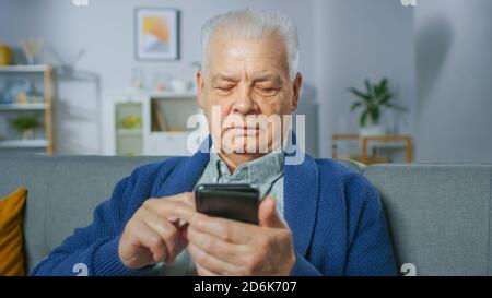 Progressiver Senior Mann sitzt in seinem Wohnzimmer leicht Smartphone verwendet, tut berührende Gesten und fühlt sich sehr wohl mit neuen Technologien. Stockfoto