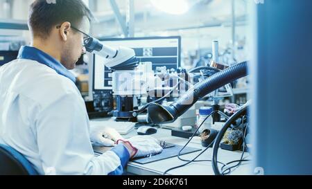 Arbeiter in der Elektronikfabrik in White Work Coat inspiziert eine Leiterplatte über ein digitales Mikroskop. High-Tech-Fabrik. Stockfoto