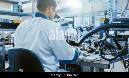 Rückansicht Aufnahme eines Arbeiters in der Elektronikfabrik in weißem Arbeitsmantel inspiziert eine Leiterplatte durch ein digitales Mikroskop. High-Tech-Fabrik Stockfoto