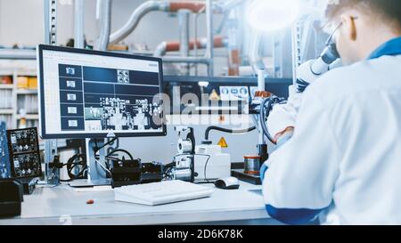 Arbeiter in der Elektronikfabrik in weißer Arbeitsschicht inspiziert eine Leiterplatte auf einem Computerbildschirm, der mit einem digitalen Mikroskop verbunden ist. Hoch Stockfoto