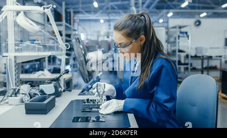 Young Female Blue and White Work Coat verwendet Zange, um gedruckte Leiterplatte für Smartphone zu montieren. Arbeiter in der Elektronikfabrik in einer High-Tech Stockfoto