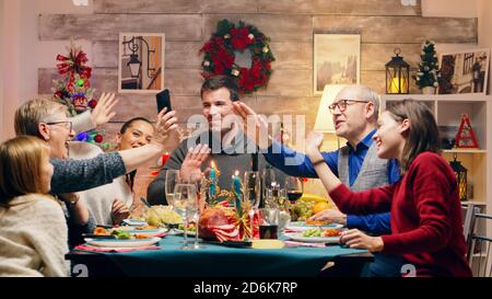 Große Familie winkt während eines Videoanrufs bei der weihnachtsfeier. Weihnachtstradition. Traditionelles festliches weihnachtsessen in einer Familie mit mehreren Generationen. Genießen Sie das Weihnachtsessen im dekorierten Zimmer. Großes Familientreffen Stockfoto