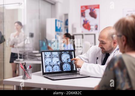 Hirnchirurg zeigt auf CT-Scan auf Laptop im Krankenhaus Büro Diskussion mit älteren Patienten über Intervention. Krankenschwester hält das Röntgenbild im Hintergrund. Junge kranke Frau im Klinikkorridor. Stockfoto